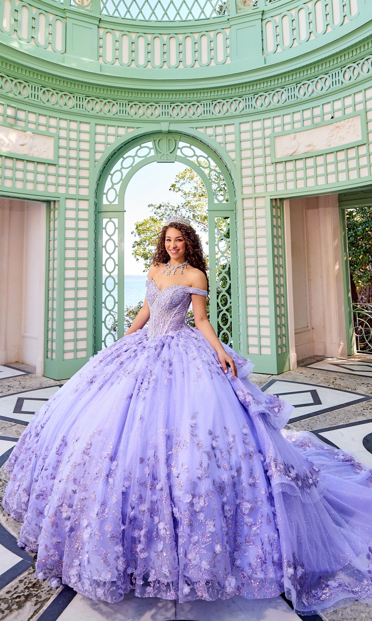 A woman in the Princesa PR30131 Off-Shoulder Quinceanera Dress with 3D floral appliques and a corset bodice stands in an ornate green and white open pavilion.